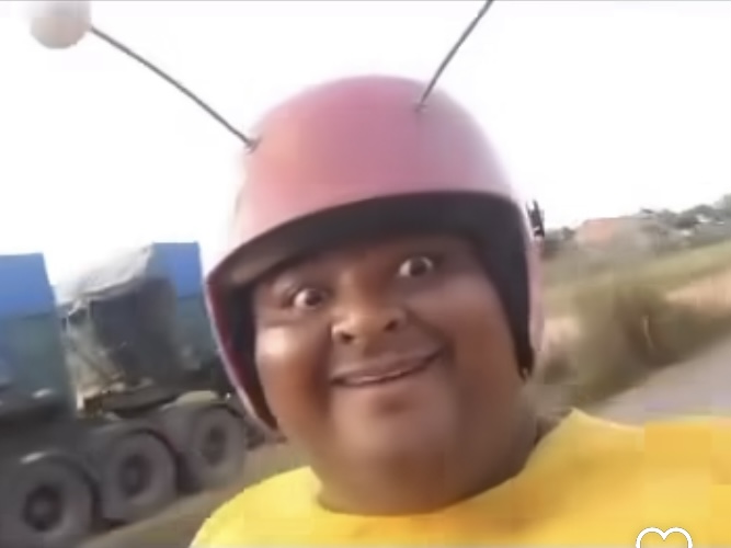 Um homem sorridente usa um capacete rosa com antenas, assemelhando-se a um personagem de inseto.